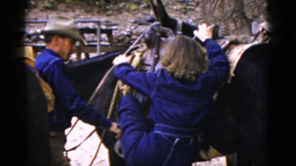 A man in Western clothing helps a woman in blue mount a saddled horse during a 1952 Arizona outdoor outing.