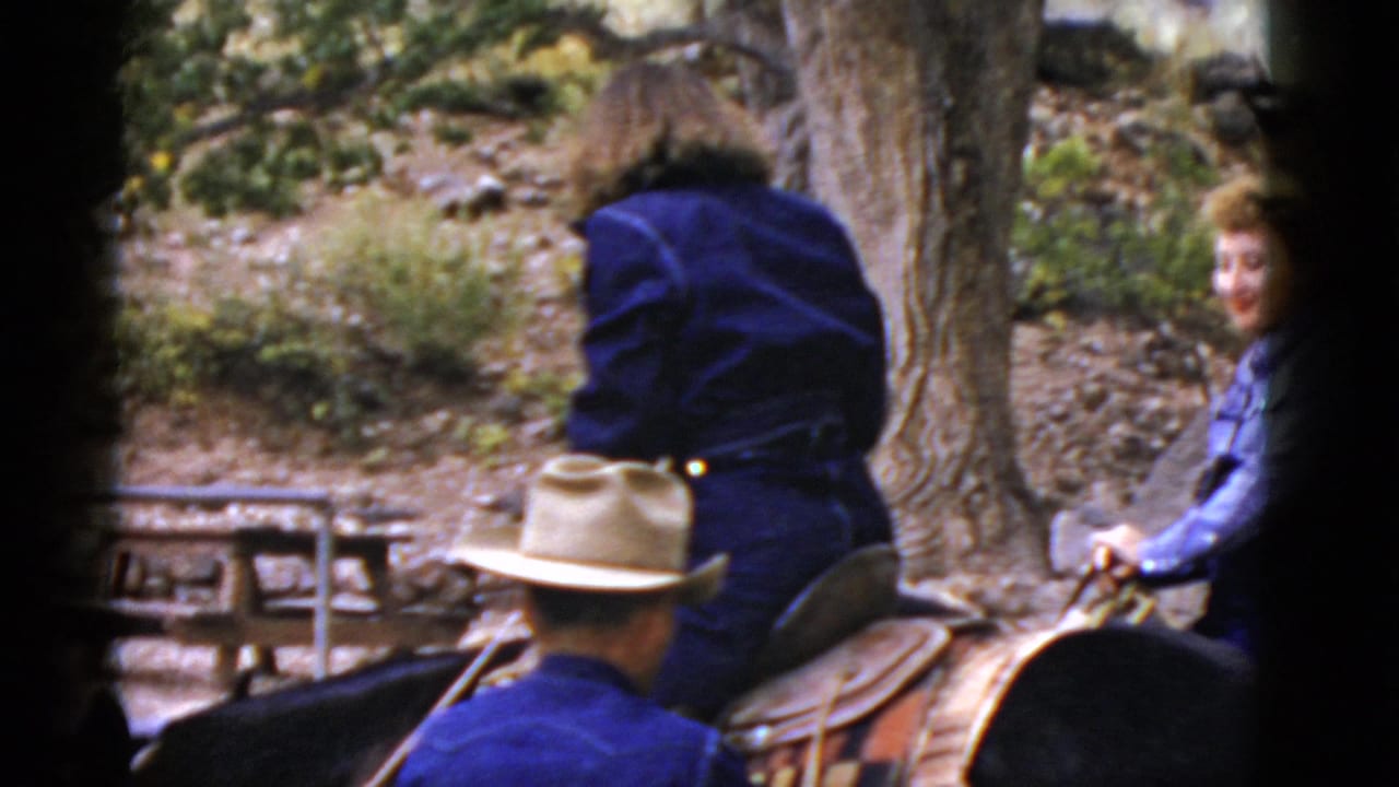 Riders and horses near trees, with a man in a cowboy hat beside a saddled horse.