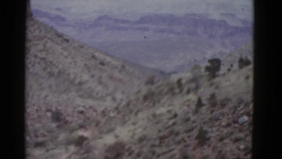1952 Arizona 8mm frame showing a rocky desert slope, scattered scrub, and distant layered mountains under a hazy sky.