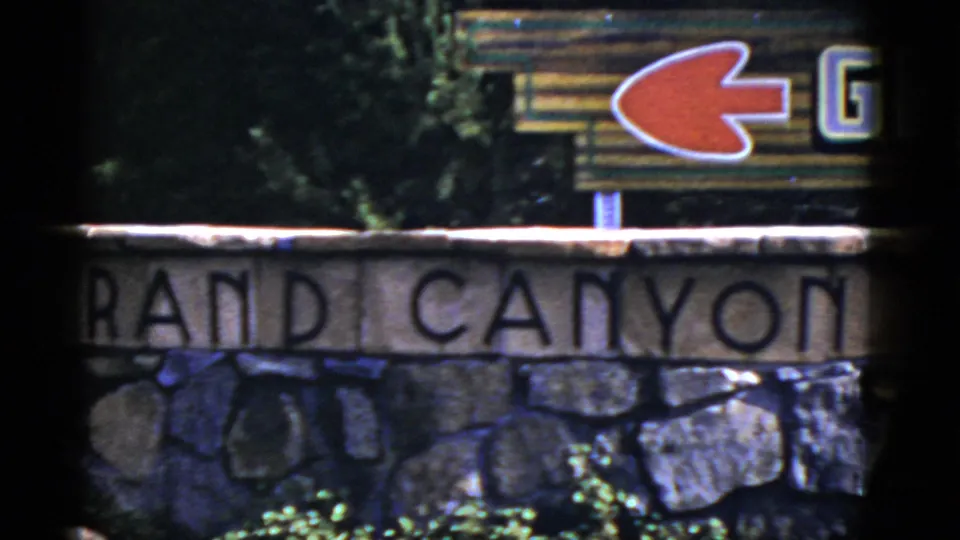 1952 Arizona 8mm frame showing carved Grand Canyon lettering, stonework, trees, and a red left-pointing arrow sign.