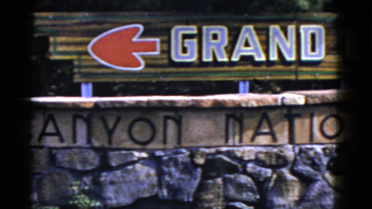 1952 Arizona home-movie frame showing part of the Grand Canyon National Park entrance sign, red arrow, wood lettering, and stone base.
