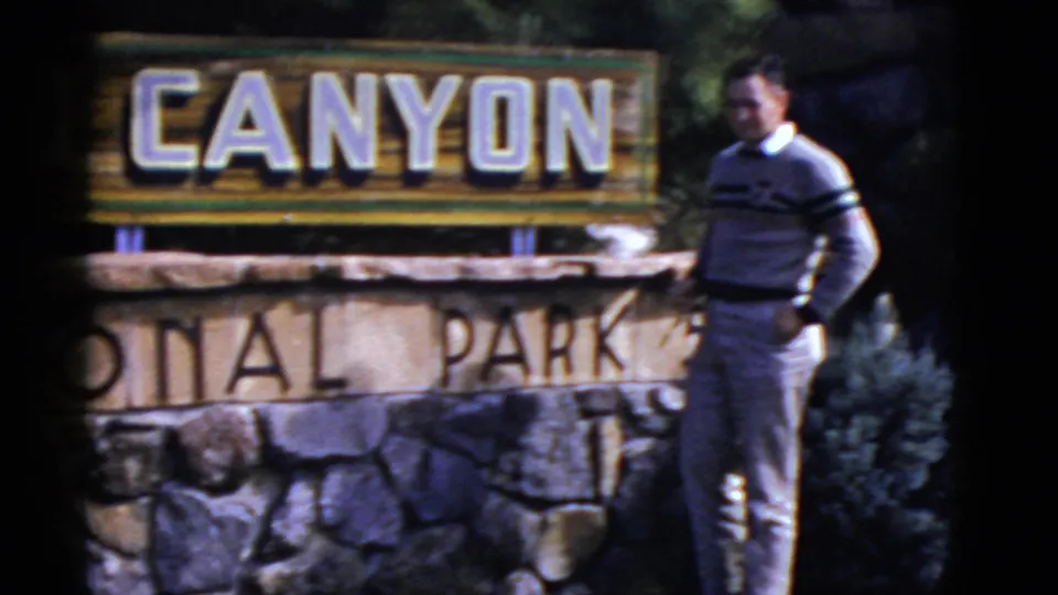 1952 Arizona frame showing an adult man in a striped sweater standing next to the Grand Canyon National Park entrance sign.