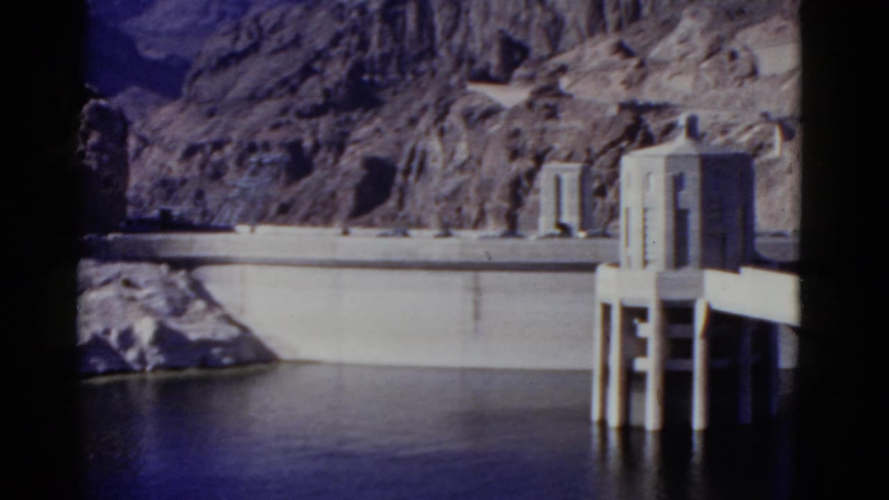Hoover Dam wall and intake tower seen across still water with rocky cliffs behind.