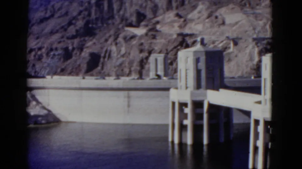 1952 Hoover Dam footage showing a concrete intake tower near the dam wall, with calm water below and desert rock cliffs beyond.