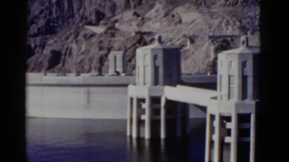 1952 8mm frame of Hoover Dam intake towers linked by a concrete walkway, with the dam wall, water, and rocky hillside visible.