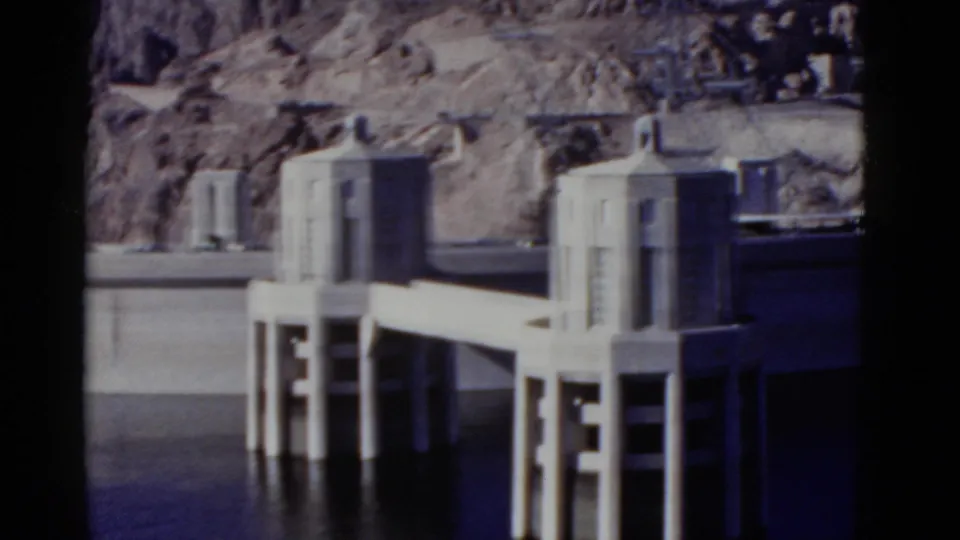 Final 1952 Hoover Dam frame with multiple intake towers, a concrete walkway, calm water, dam wall, and rugged canyon rock in view.
