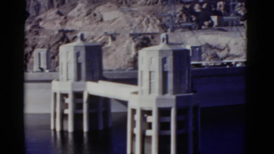 1952 California 8mm frame showing concrete dam intake towers, a deck, vertical supports, reservoir water, and rocky desert terrain.