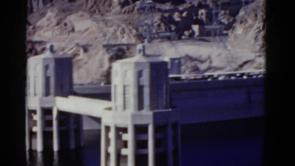 1952 California 8mm frame of a concrete dam structure with twin towers, elevated deck, dark water, and rugged desert cliffs.