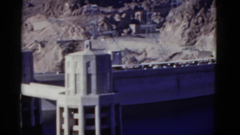 1952 California 8mm frame showing a concrete dam tower in the foreground, a roadway or deck, reservoir water, and rocky desert hillside.