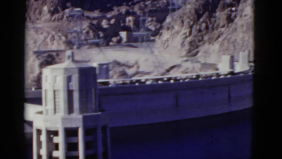 1952 California 8mm frame showing a dam wall, concrete intake tower, dark reservoir water, and rugged desert rock formations.