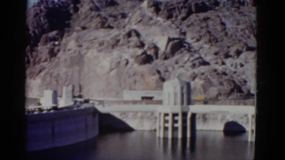 1952 California 8mm frame showing the dam edge at left, intake towers at right, still water, and rugged rock formations behind.