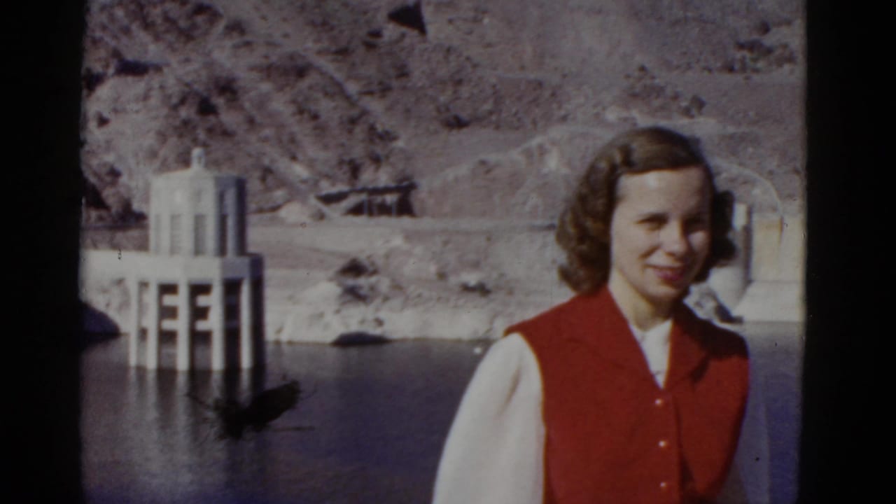 In 1952 California home-movie footage, a young woman in a red vest and white blouse poses near a dam or reservoir with canyon slopes behind her.