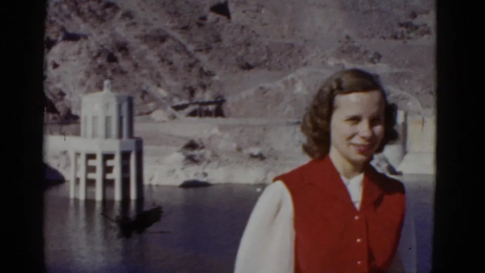 In 1952 California home-movie footage, a young woman in a red vest and white blouse poses near a dam or reservoir with canyon slopes behind her.
