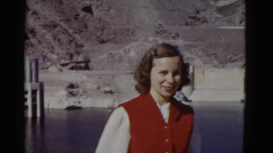 The woman remains posed near the California dam setting, smiling toward the camera as reservoir water and concrete works fill the background.