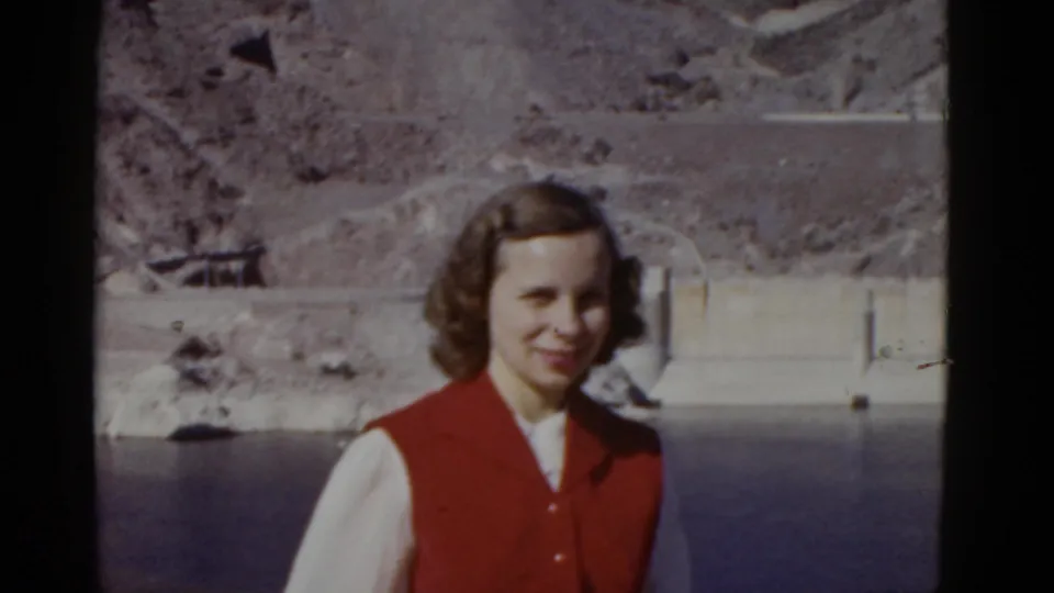 A closer 1952 California frame shows the woman in period clothing standing before reservoir water, concrete dam features, and rocky canyon terrain.