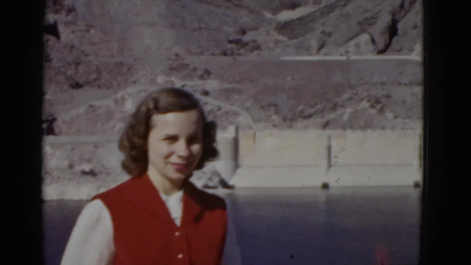 The final frame keeps the woman centered against the California dam or reservoir backdrop, with water, concrete, and rocky slopes visible.