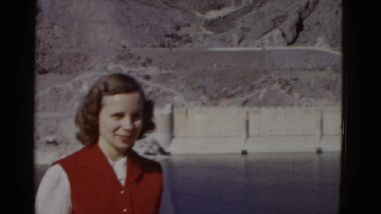 In 1952 California home-movie footage, a young woman in a red vest smiles beside dark water with a concrete dam and arid hillside behind her.