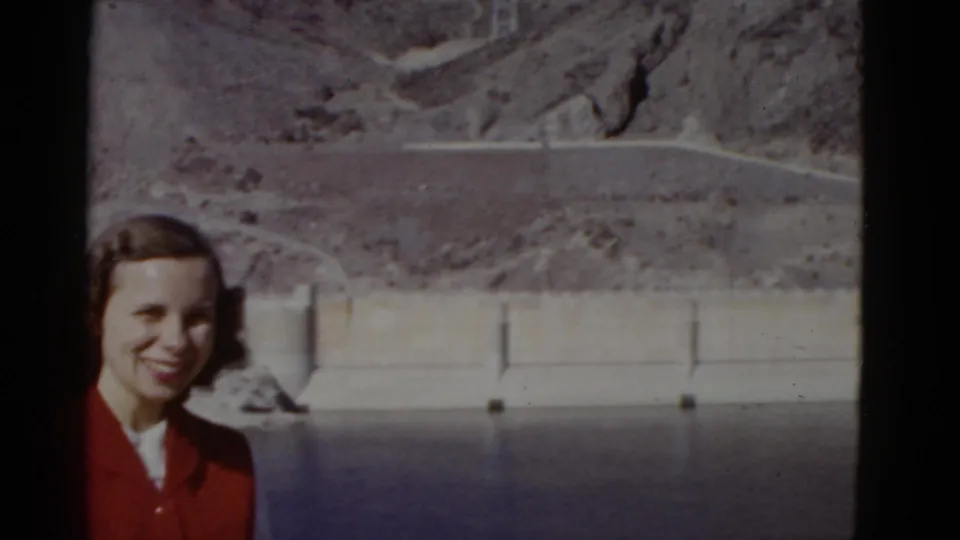 The woman smiles near the edge of the frame, with the reservoir, concrete dam structure, and dry mountain slope visible behind her.