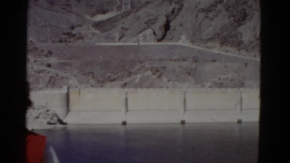 The camera drifts left of the subject, leaving only part of her red vest visible while the dam, water, and rocky California landscape fill the frame.