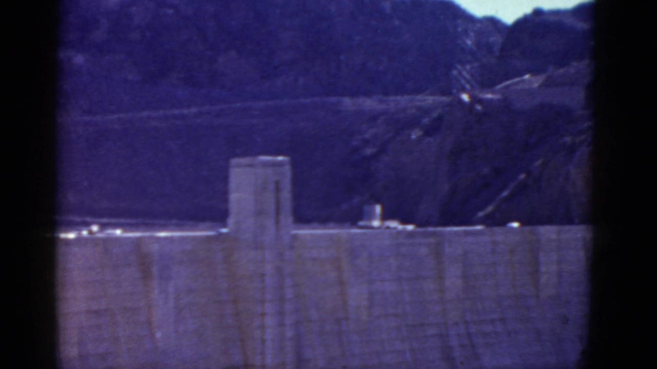 1952 8mm frame of Hoover Dam from a distance, showing the concrete face, crest area, rugged hills, and soft blue-purple film color.