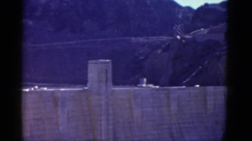 1952 8mm frame of Hoover Dam from a distance, showing the concrete face, crest area, rugged hills, and soft blue-purple film color.