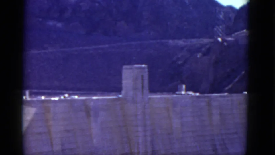 Distant 1952 view of Hoover Dam, with the dam face, crest roadway, small vehicles, and dark rocky hills softened by aged 8mm color.