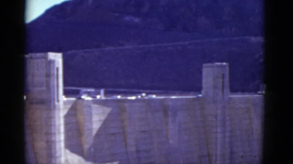 End frame of the 1952 Hoover Dam view, with tower structures, crest traffic, concrete wall, and rugged canyon hills in aged 8mm color.