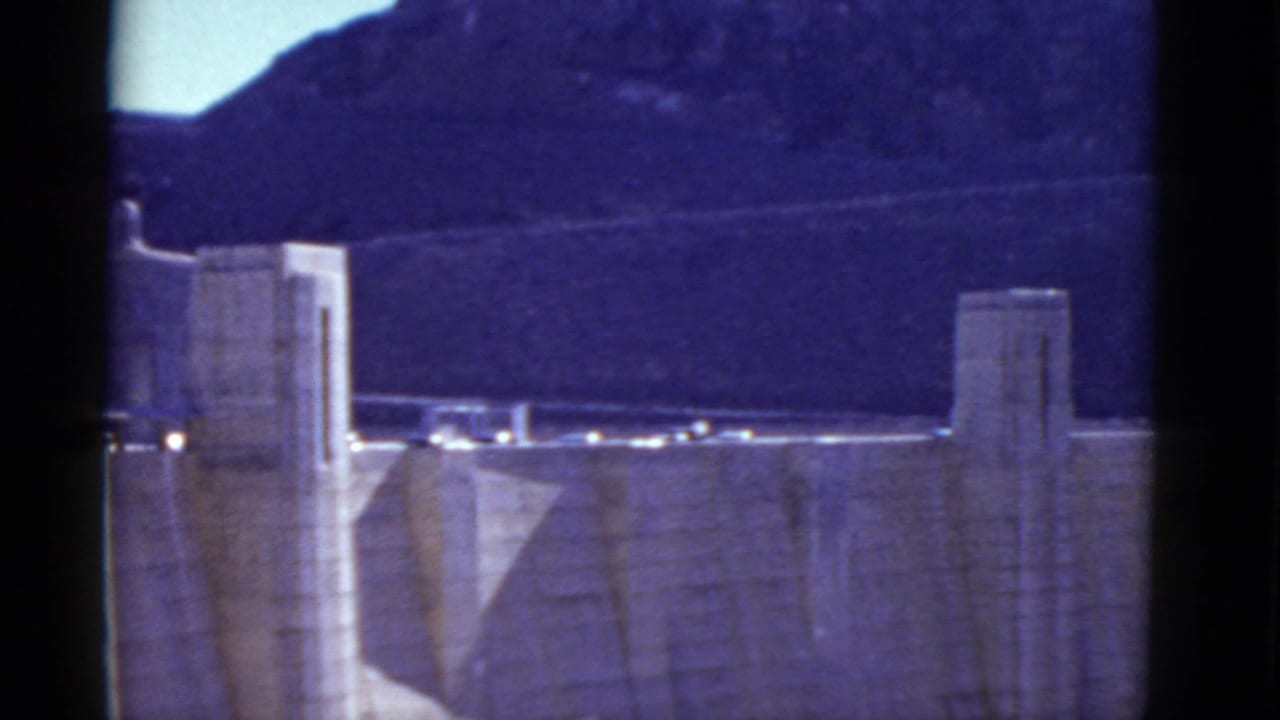Concrete dam towers and roadway seen against dark mountains and bright sky.