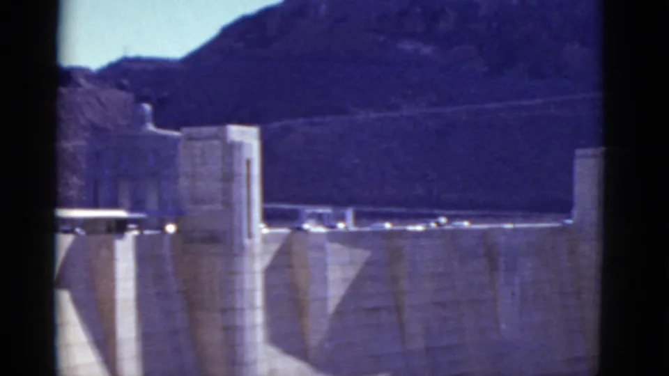 1952 8mm footage shows a Hoover Dam tower and roadway with small cars, set below rugged mountains and a bright sky.