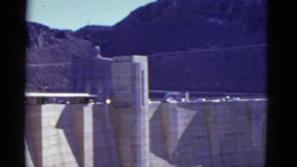 1952 8mm home-movie frame of Hoover Dam with a tower, concrete face, road deck, and rocky mountain terrain in view.