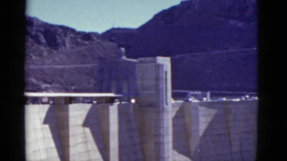 1952 8mm home-movie view of Hoover Dam's tower, concrete buttresses, crest roadway, and surrounding desert mountains.
