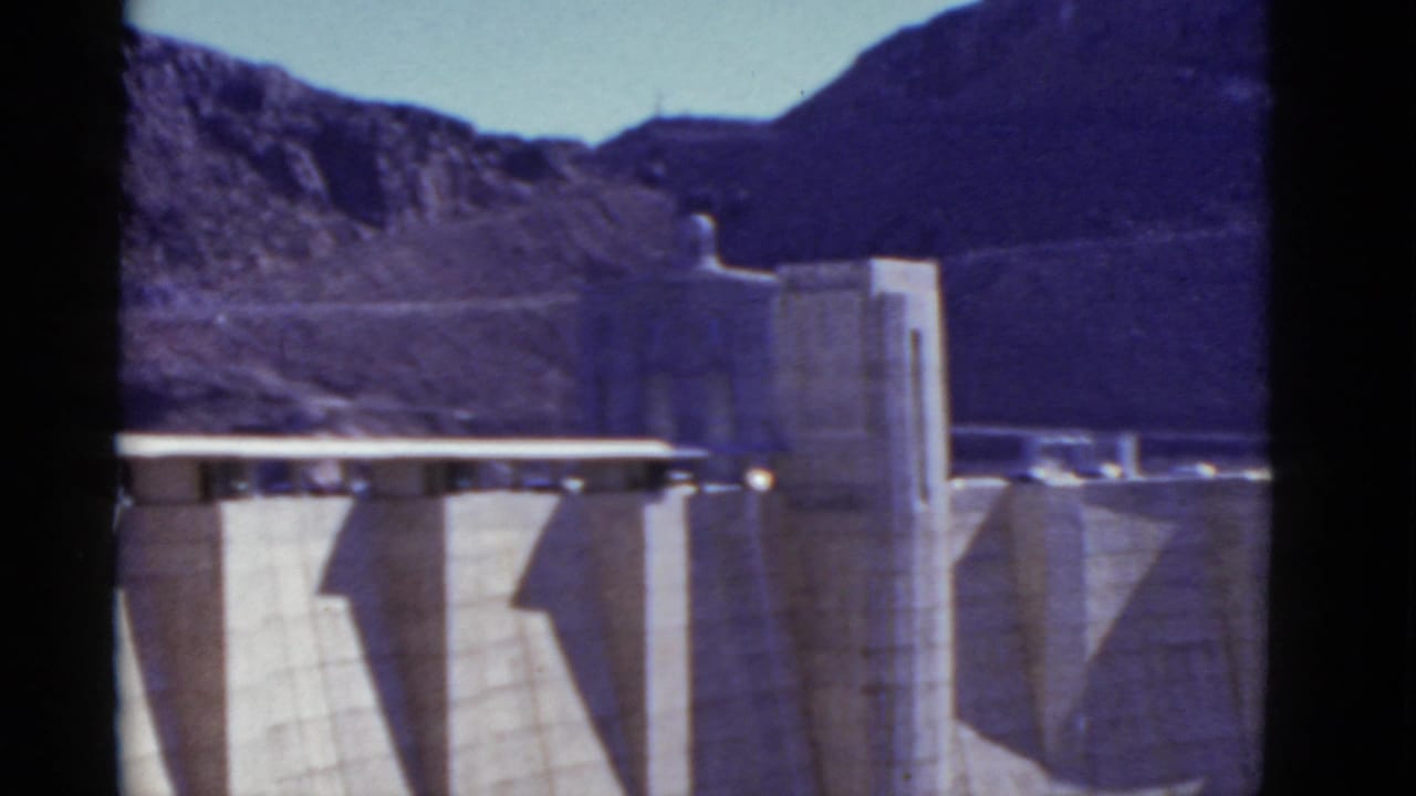 Concrete dam face and tower structures against rugged mountains and blue sky.