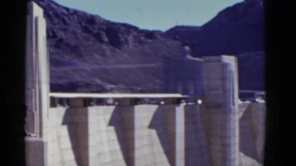 1952 California 8mm view of a concrete dam span with tall tower forms and mountain ridges behind it.