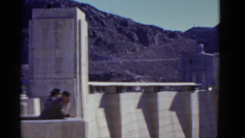 1952 California 8mm frame showing two visitors watching a concrete dam from an overlook, with mountains in the distance.