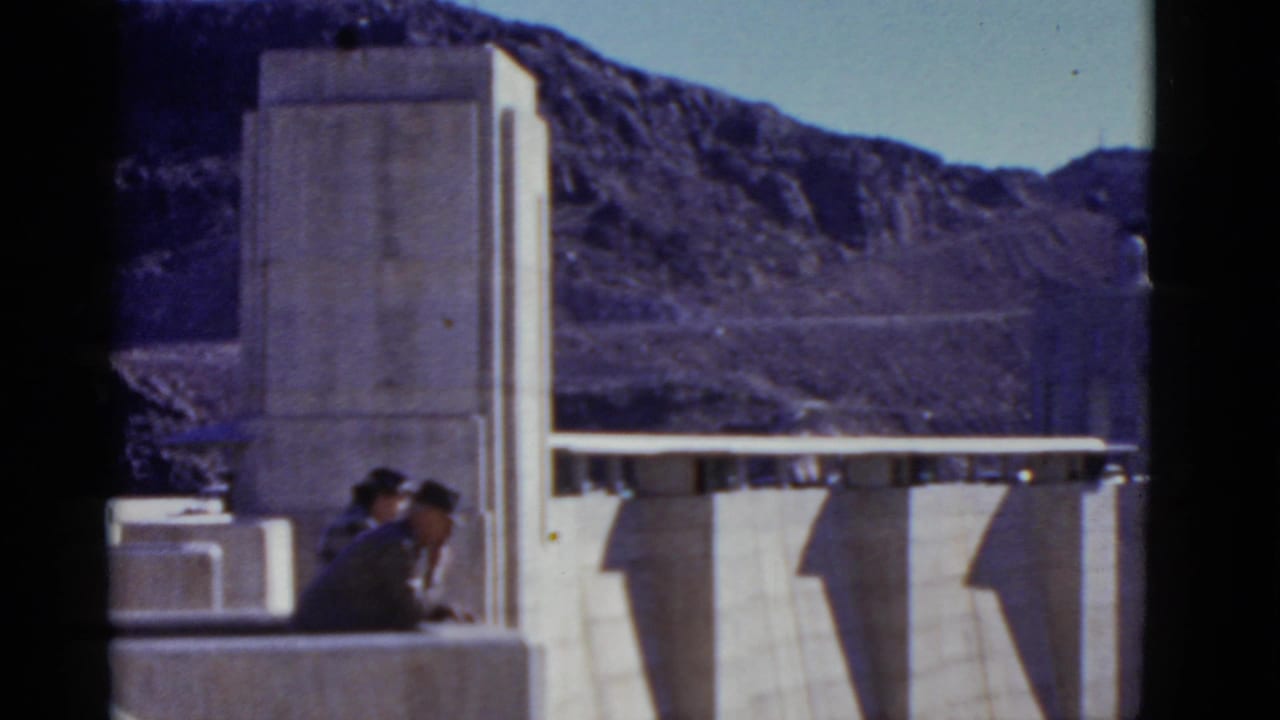1952 California 8mm frame showing two hat-wearing visitors near a concrete dam, backed by arid mountain slopes and clear sky.