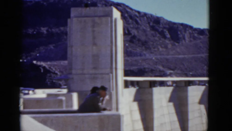 1952 California home-movie frame of two visitors seated near a tall concrete dam structure under a clear sky.