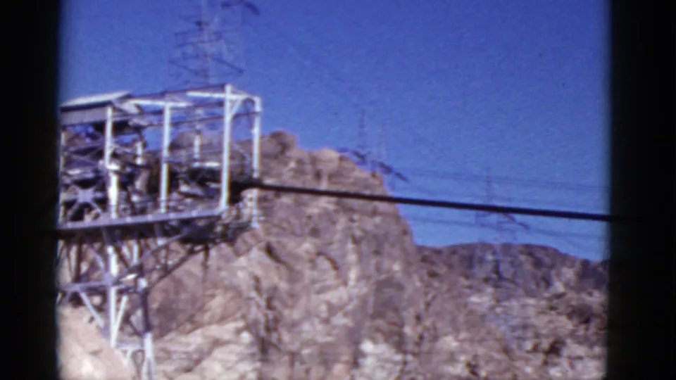 1952 California 8mm frame showing a steel transmission tower on a rocky outcrop with power lines crossing a blue sky.