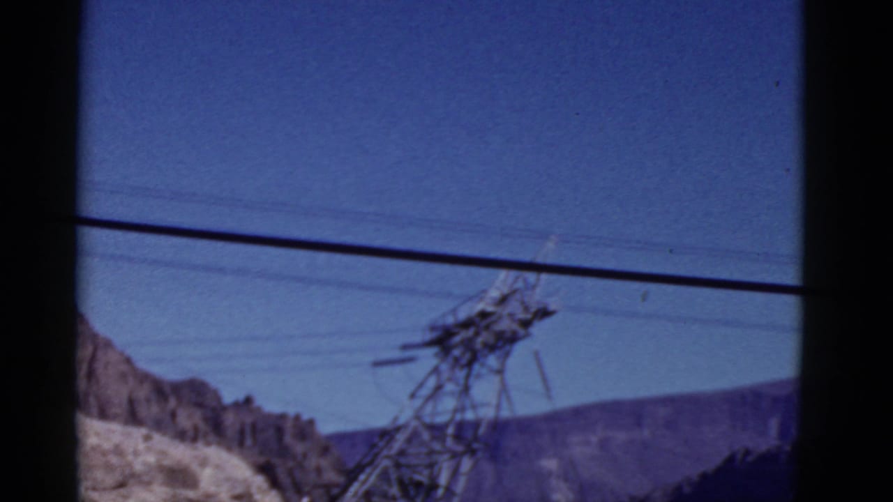 1952 California 8mm frame showing a steel transmission tower, overhead lines, blue sky, and arid mountain slopes from a moving vehicle.
