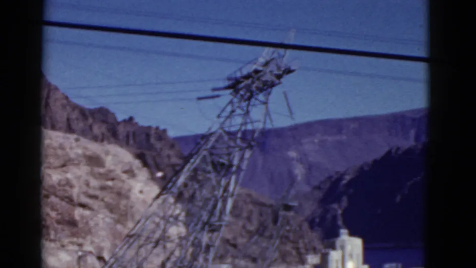 1952 California 8mm frame of a transmission tower and overhead wires, with rocky mountains and a dark valley shape behind.