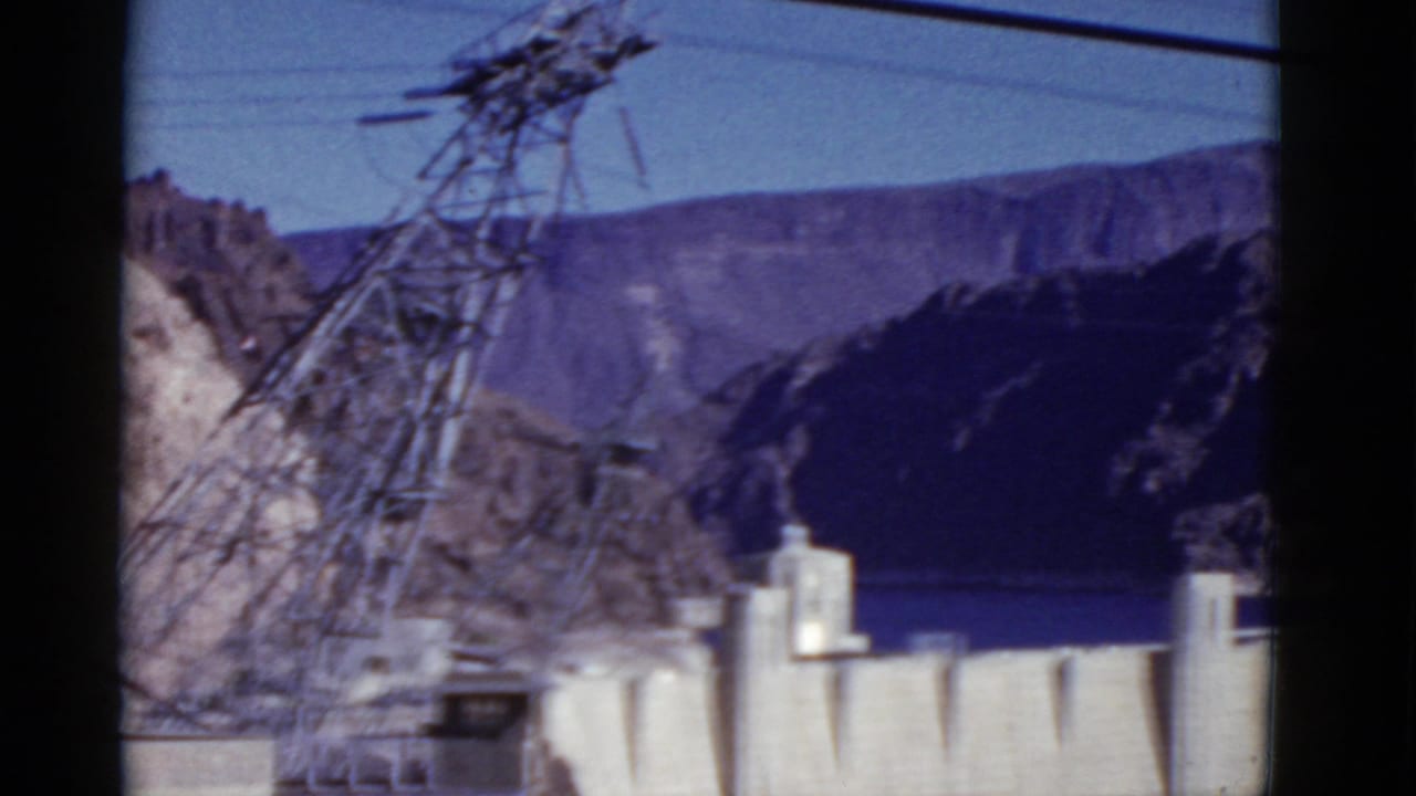 1952 8mm frame of Hoover Dam from an elevated visitor viewpoint, with a foreground transmission tower and canyon walls beyond.