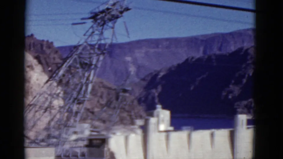 1952 8mm frame of Hoover Dam from an elevated visitor viewpoint, with a foreground transmission tower and canyon walls beyond.