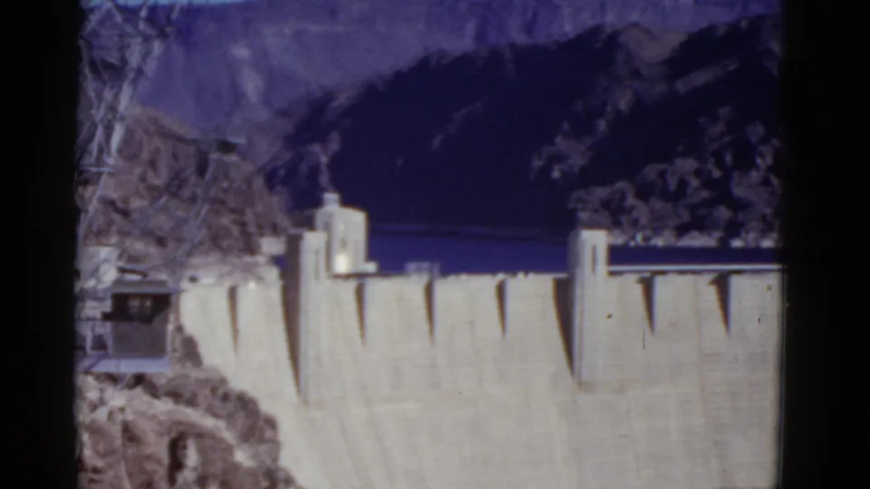 1952 8mm frame looking across Hoover Dam toward reservoir water and rugged canyon walls, with foreground branches at left.