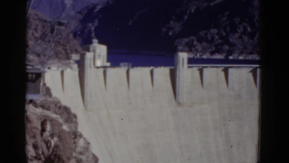 1952 home-movie frame of Hoover Dam's concrete face and towers, set against reservoir water and shadowed canyon terrain.