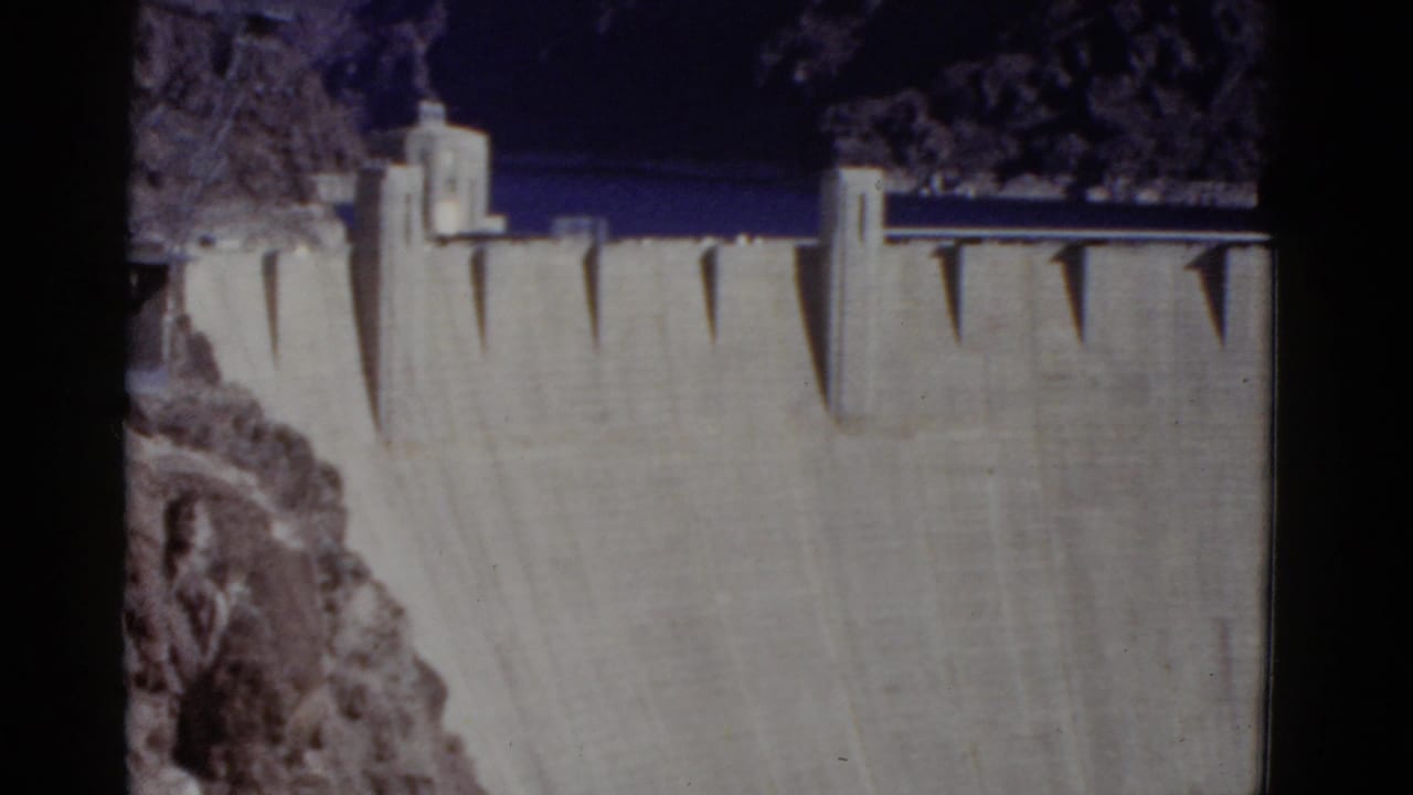 1952 8mm frame of Hoover Dam from above, showing the upper concrete face, crest structures, dark reservoir water, and rocky canyon edge.