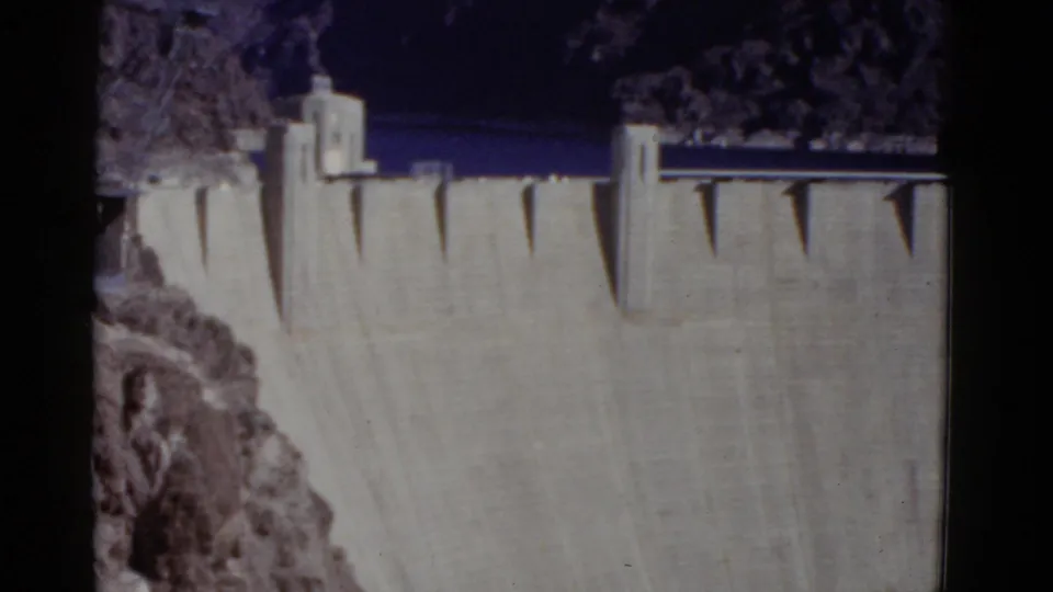 1952 8mm frame of Hoover Dam from above, showing the upper concrete face, crest structures, dark reservoir water, and rocky canyon edge.