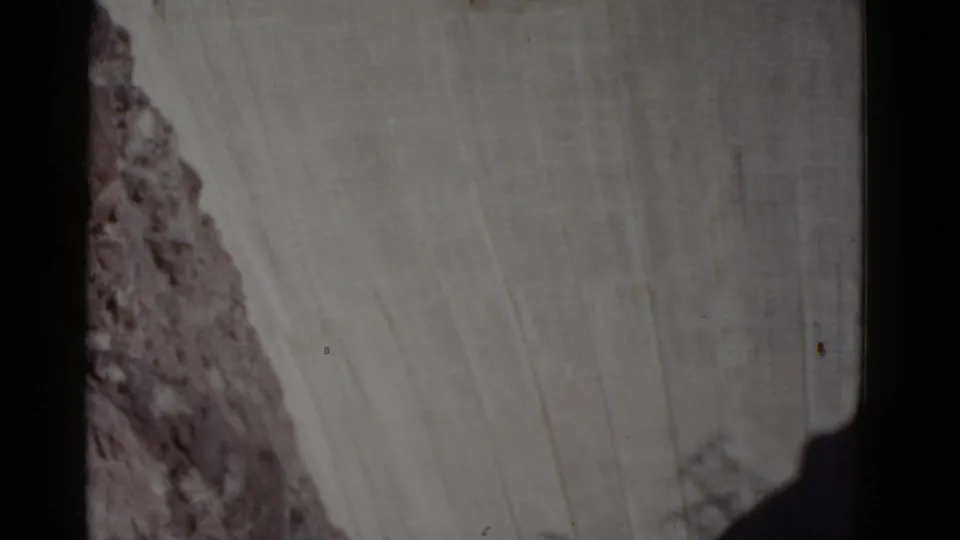 1952 8mm frame from the downward pan showing the broad concrete wall of Hoover Dam, canyon rock at left, and deep shadow near the bottom edge.