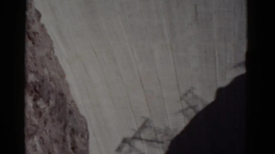 1952 8mm frame shows the dam wall descending toward a shadowed lower area, with rocky canyon terrain and a faint structural outline near the bottom.