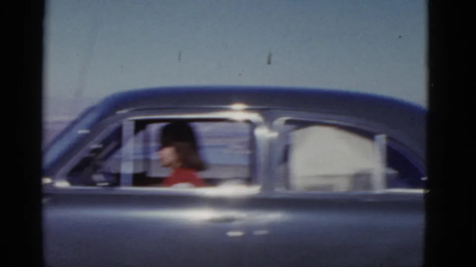 A woman wearing a red top is seen in profile through the side window of a dark mid-century sedan in 1952 California footage.