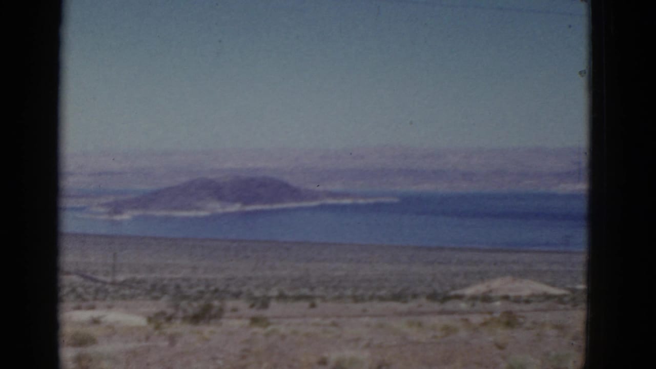 Desert foreground with low shrubs, blue water, and distant mountains under a hazy sky.