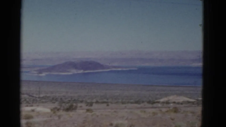 1952 California 8mm frame showing arid roadside terrain, a broad body of water, and distant mountain ridges beyond the shore.